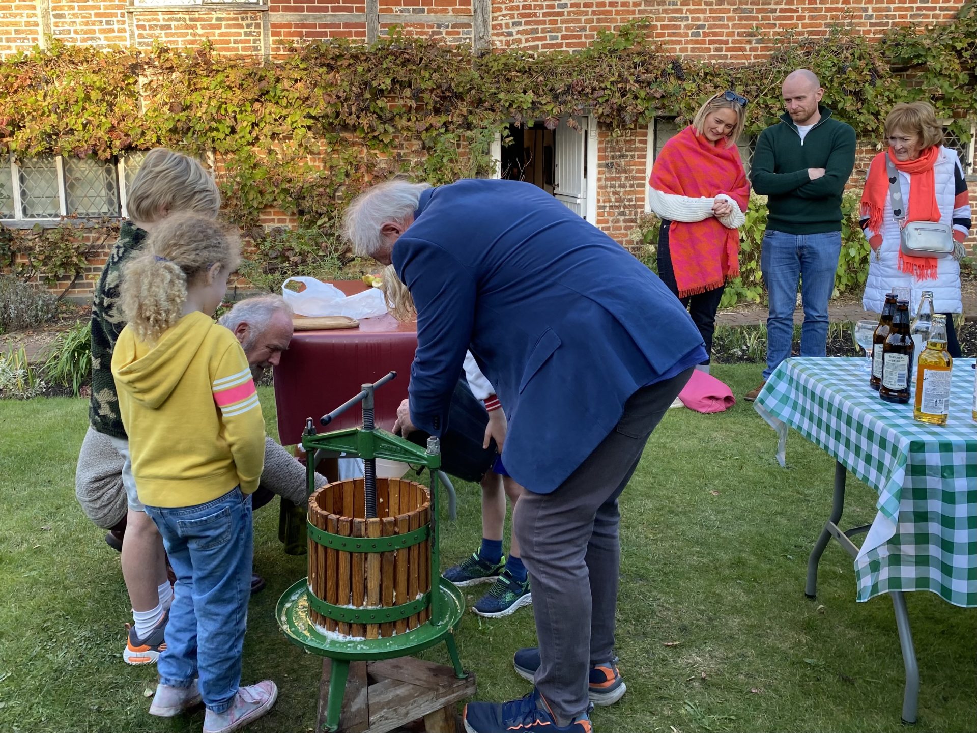 Two children and two adults are using an apple press to make juice in the autumn sunshine. Milton's Cottage is in the background, with some adults watching. Bottles of the squeezed apple juice are in the foreground of the photo.
